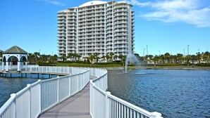 A waterfront promenade with a white bridge leading to a gazebo, a large hotel with balconies, and a fountain in the water. - Terrace @ Pelican Beach Resort 307b * Hotel accommodation * Salt Water Vacations (Destin)