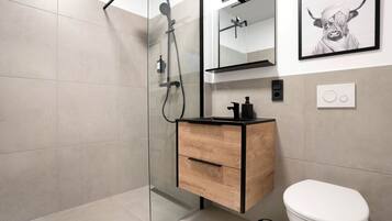 Un baño moderno con ducha envuelta en vidrio, tocador de madera, inodoro blanco y una obra de arte enmarcada en la pared.