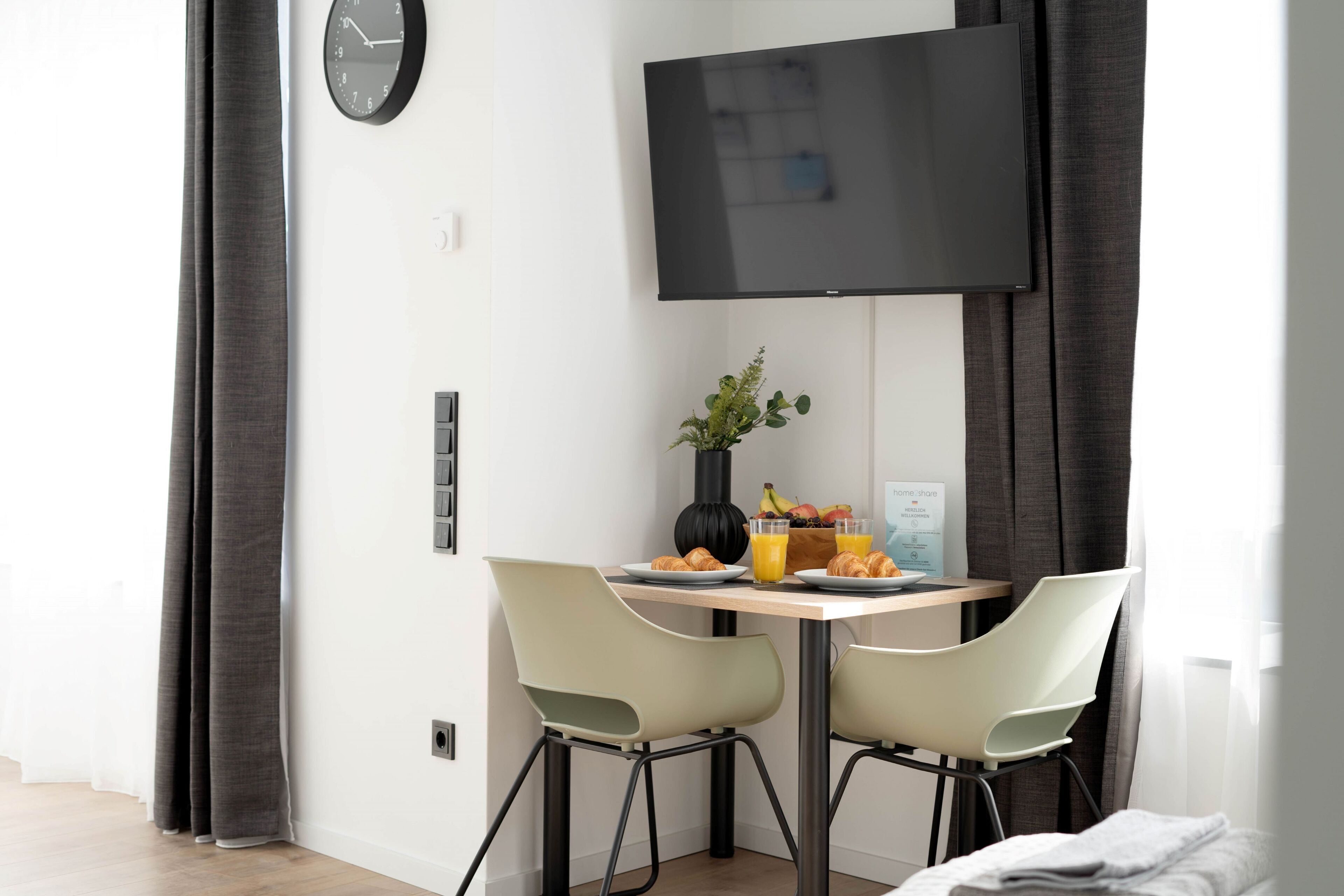 A modern hotel room with a dining area featuring two chairs, a table with breakfast items, a flat-screen TV, and a clock on the wall.