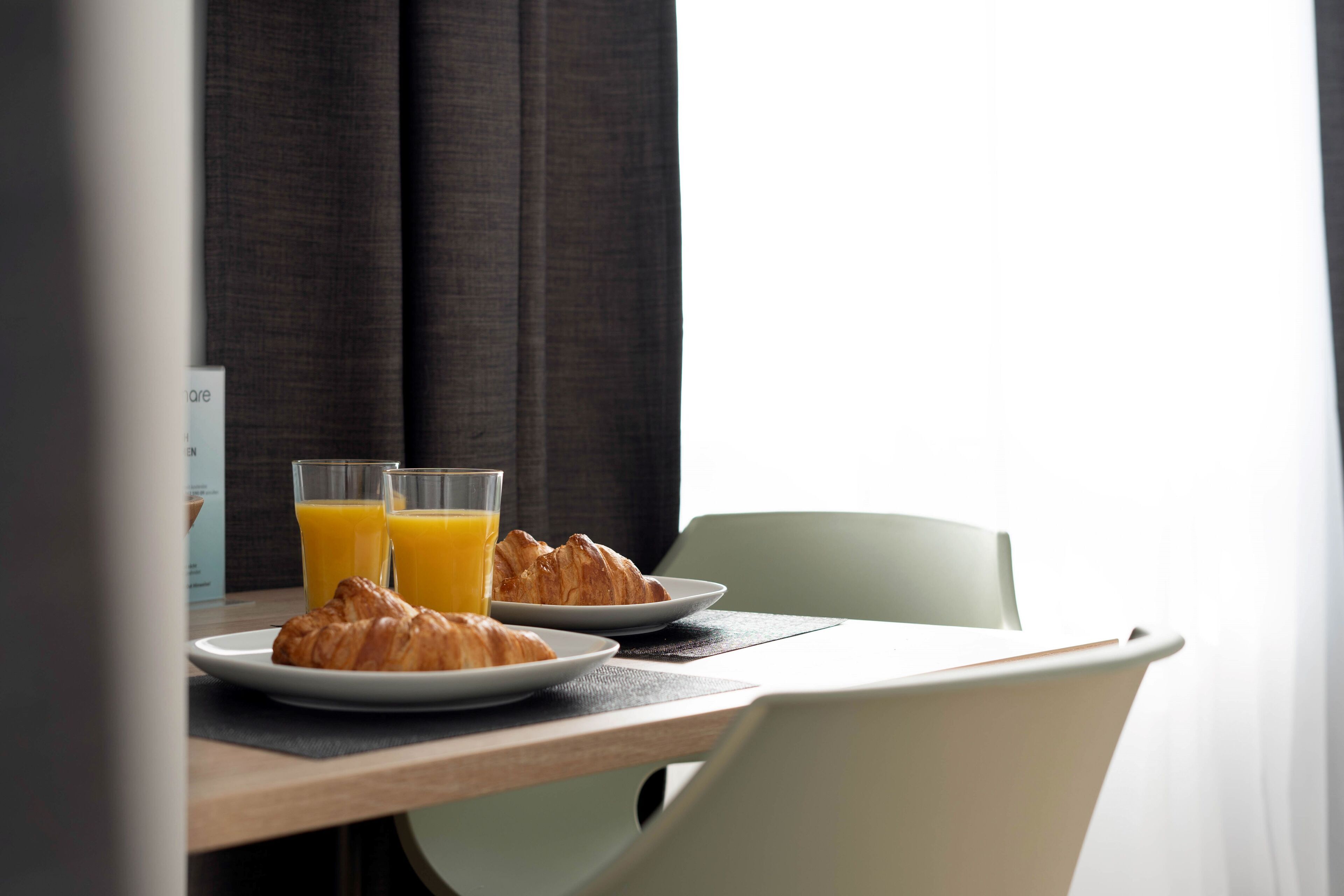 A breakfast setup with croissants and orange juice on a table, a chair, and a window with curtains.