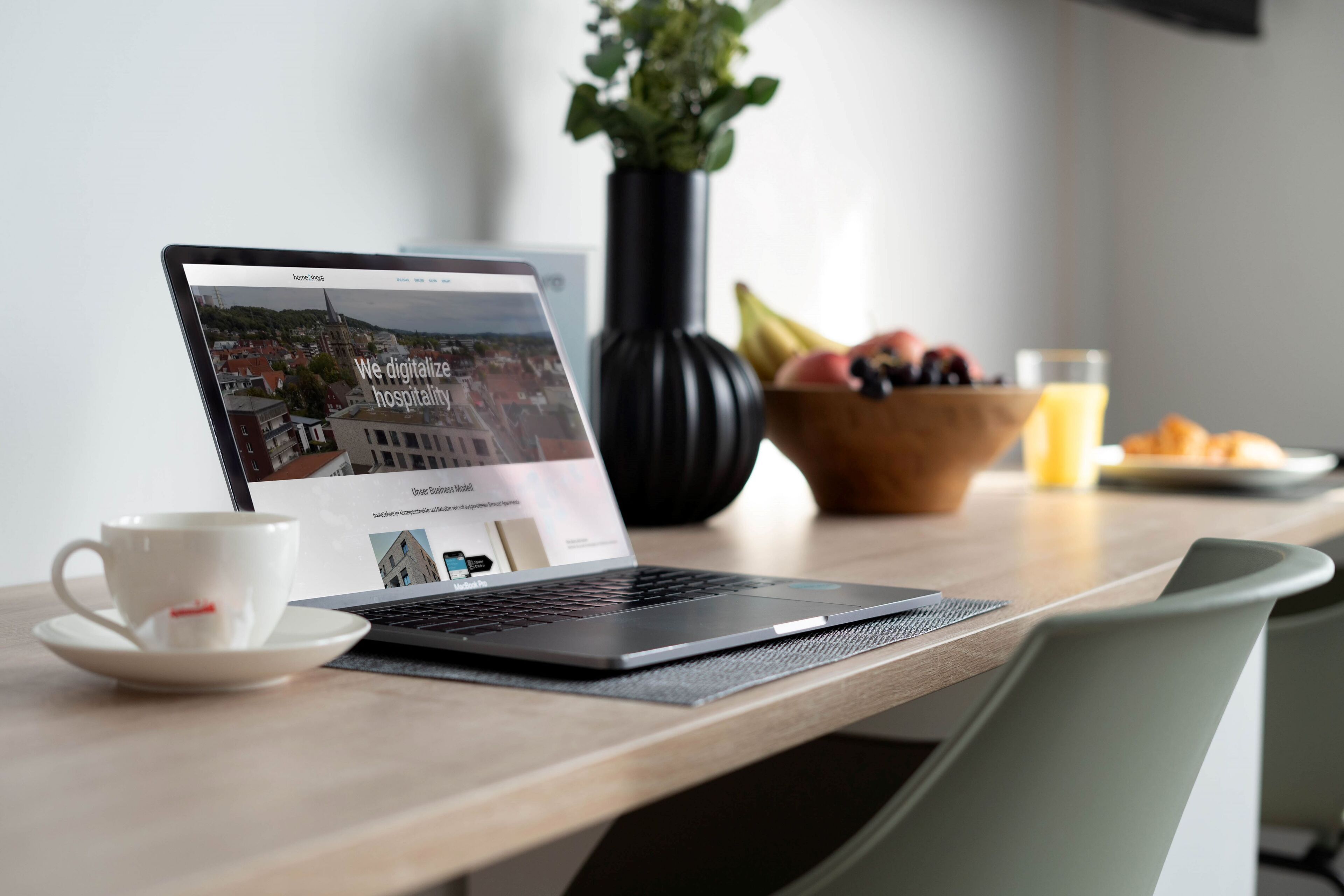 A laptop on a wooden table with a cup and saucer, a bowl of fruit, and a glass of juice.