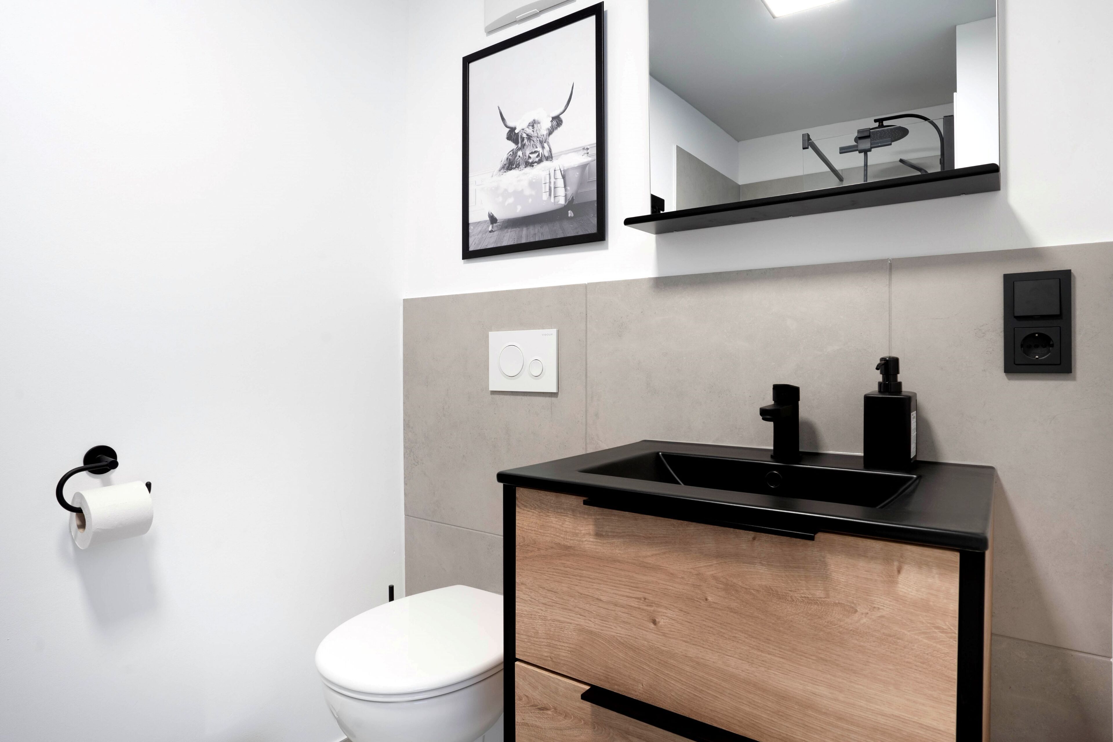 Un baño moderno con inodoro blanco, lavamanos negro con gabinete de madera, un cuadro enmarcado en la pared y un estante sobre el lavamanos.