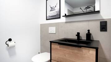 Un baño moderno con inodoro blanco, lavamanos negro con gabinete de madera, un cuadro enmarcado en la pared y un estante sobre el lavamanos.