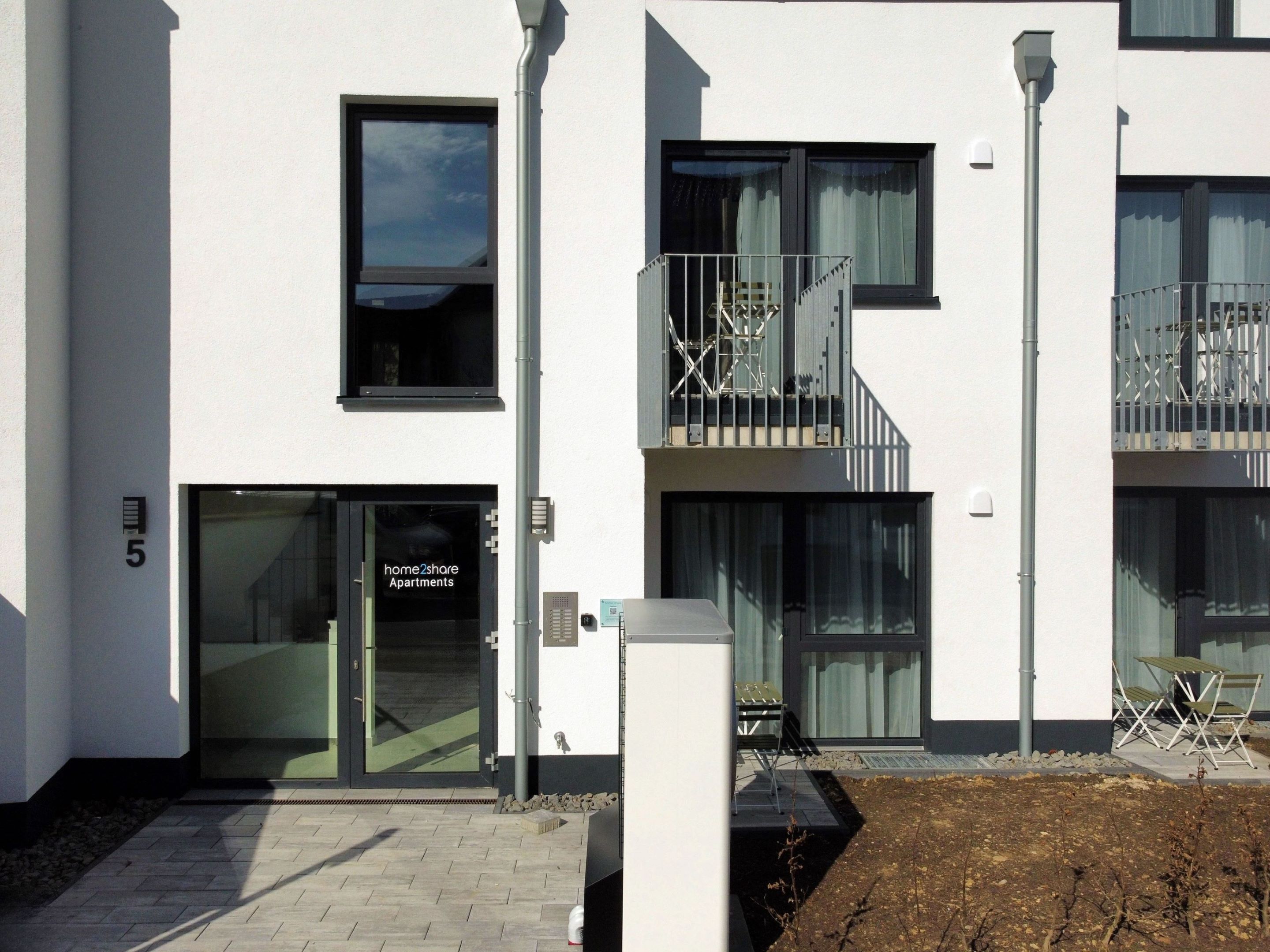 A modern apartment building with a glass entrance, a balcony with a metal railing, and a sign indicating 'home2share apartments'.