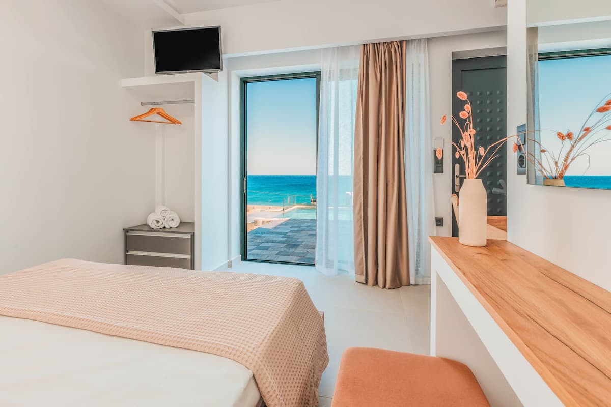 Een hotelkamer met een bed, een houten nachtkastje, een aan de muur bevestigde televisie, uitzicht op de oceaan en een deur naar een balkon.