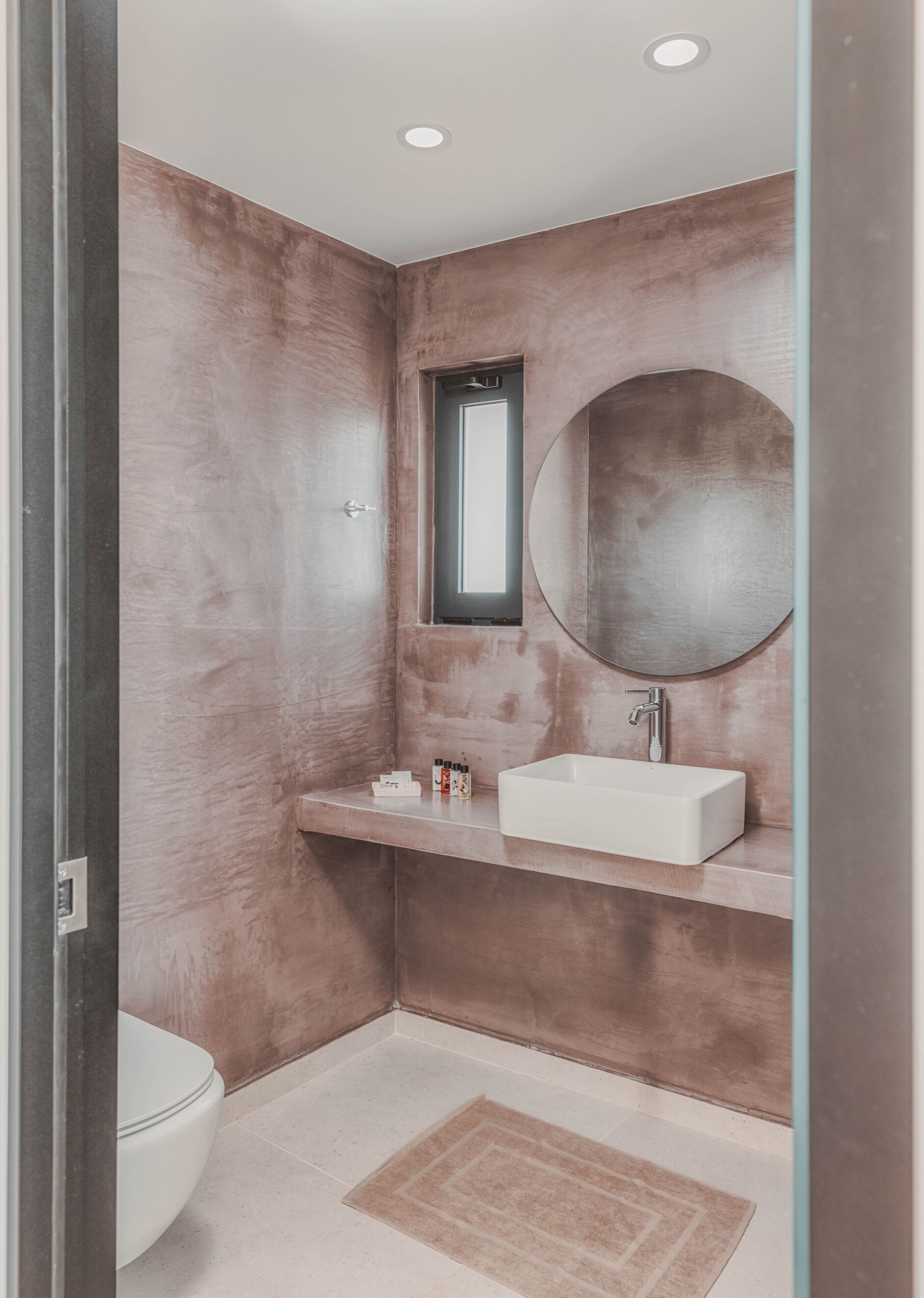 A modern bathroom with a rectangular sink, a round mirror, and a small window.