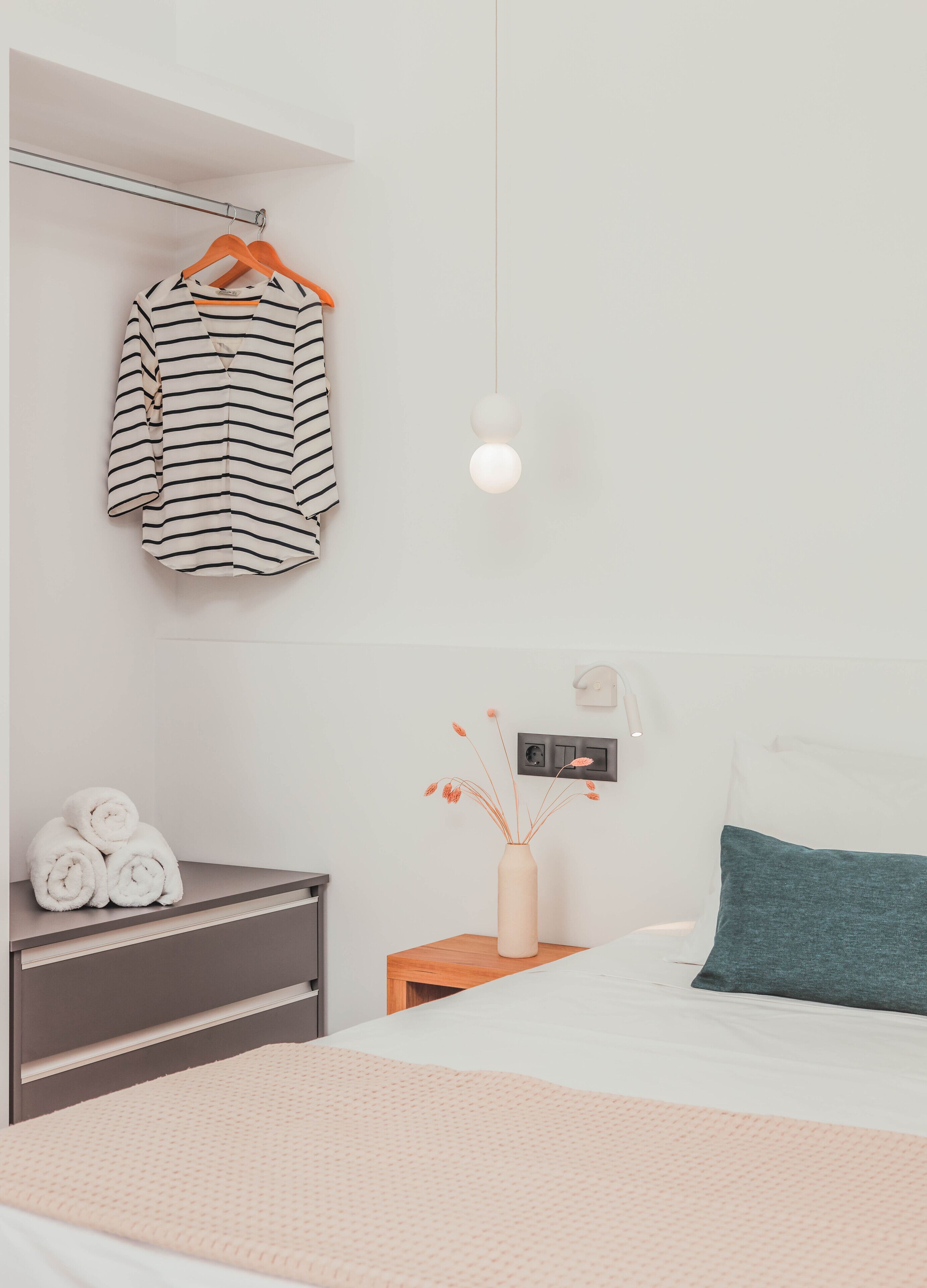 A modern bedroom with a neatly made bed, a bedside table with towels, a hanging light fixture, and a bedside lamp.