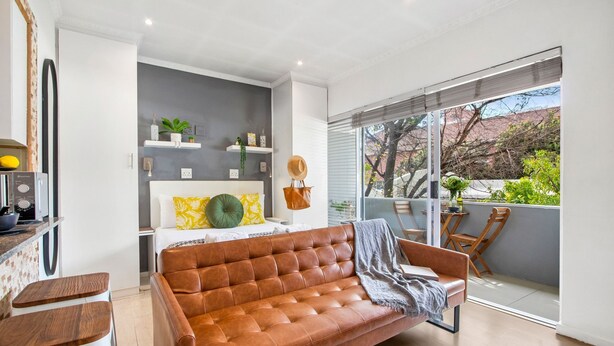 A modern living room with a brown leather sofa, a dining area with a wooden table, and a balcony with a view of trees and buildings.