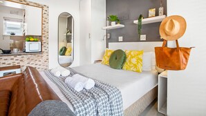 A modern hotel room with a bed, a sofa, a mirror, a small kitchen area, and a decorative hat on a stand.