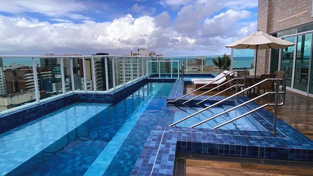 Une piscine à débordement située sur le toit, avec une terrasse en bois, des chaises longues et une balustrade en verre.