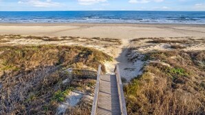 Een houten looppad dat naar een zandstrand met duinen en schaarse begroeiing leidt.