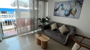 A modern living room with a grey sofa, a wooden coffee table, and a wooden rocking chair. There is a large window with a view of a balcony and a building outside. - Rodadero Excalibur 8c (Santa Marta)