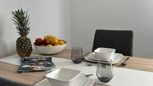 Ein Esstisch ist gedeckt mit weißen Tellern, Schüsseln, Besteck, einem Glas und einer Zeitschrift. Auf dem Tisch befinden sich eine Ananas und eine Obstschale.