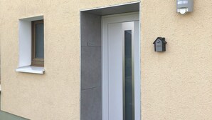 Ein Gebäudeeingang mit Tür, kleinem Fenster, Wandleuchte und Mülleimer.