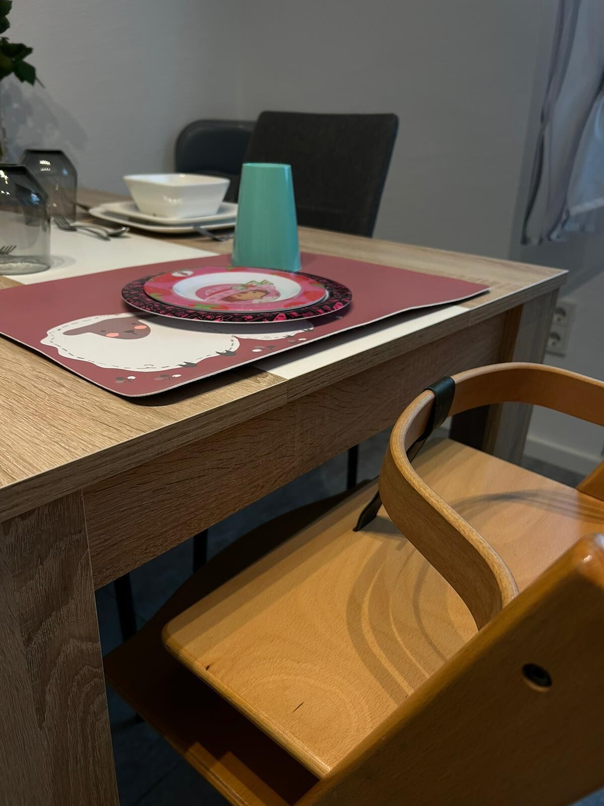 A wooden dining table with a placemat featuring a cartoon character, a teal cup, and a white bowl.