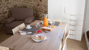 Una mesa de desayuno con pan, fruta y un vaso de jugo de naranja. En el fondo, se aprecia una pared de piedra y un armario blanco.