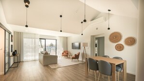 Un salon spacieux doté d’un plancher en bois, d’un canapé, d’une table basse et d’un coin repas avec des chaises et une table. On y trouve également des luminaires suspendus, un téléviseur à écran plat et une grande fenêtre avec des rideaux.