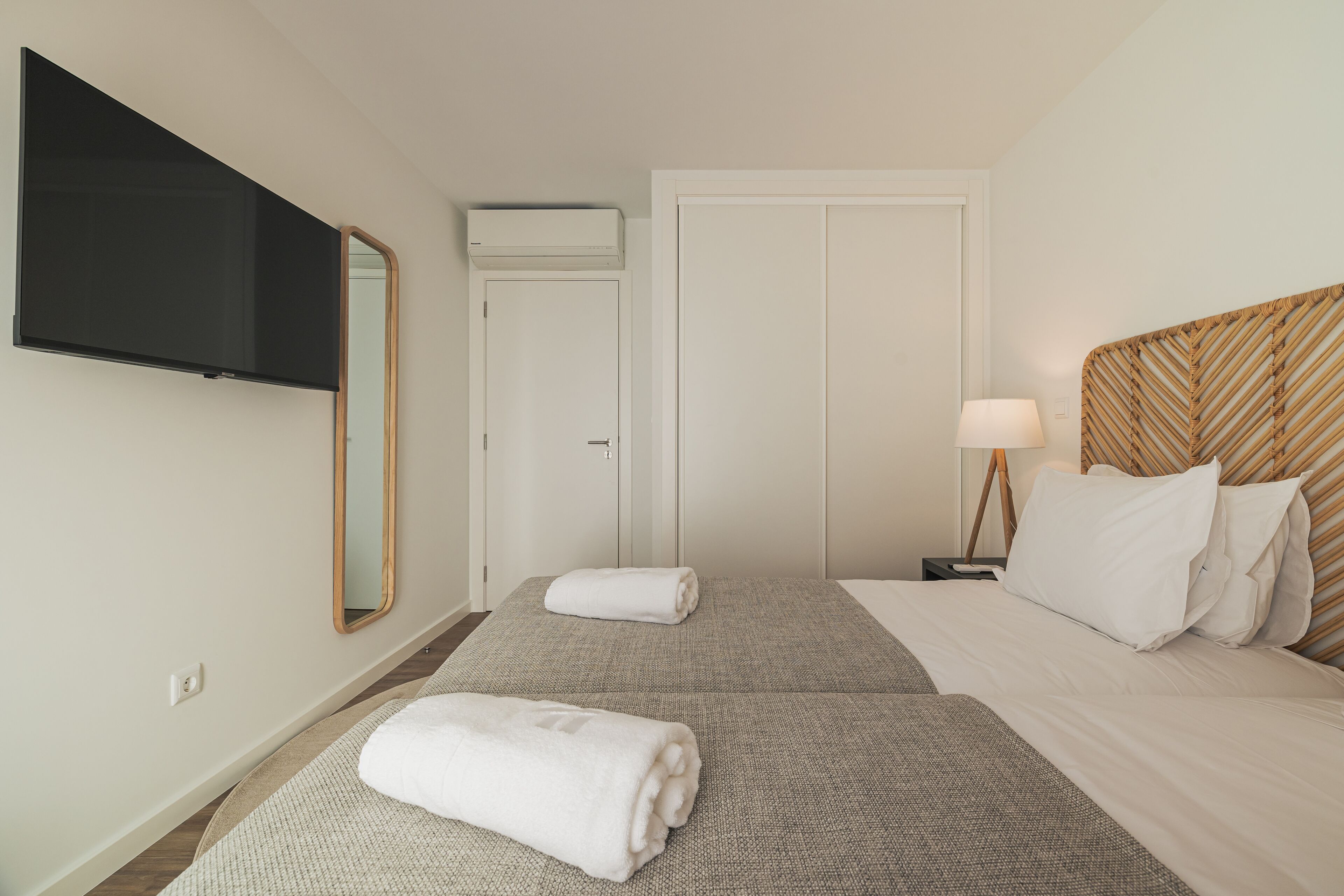 A hotel room with two beds, a television mounted on the wall, a door, a nightstand with a lamp, and a mirror.