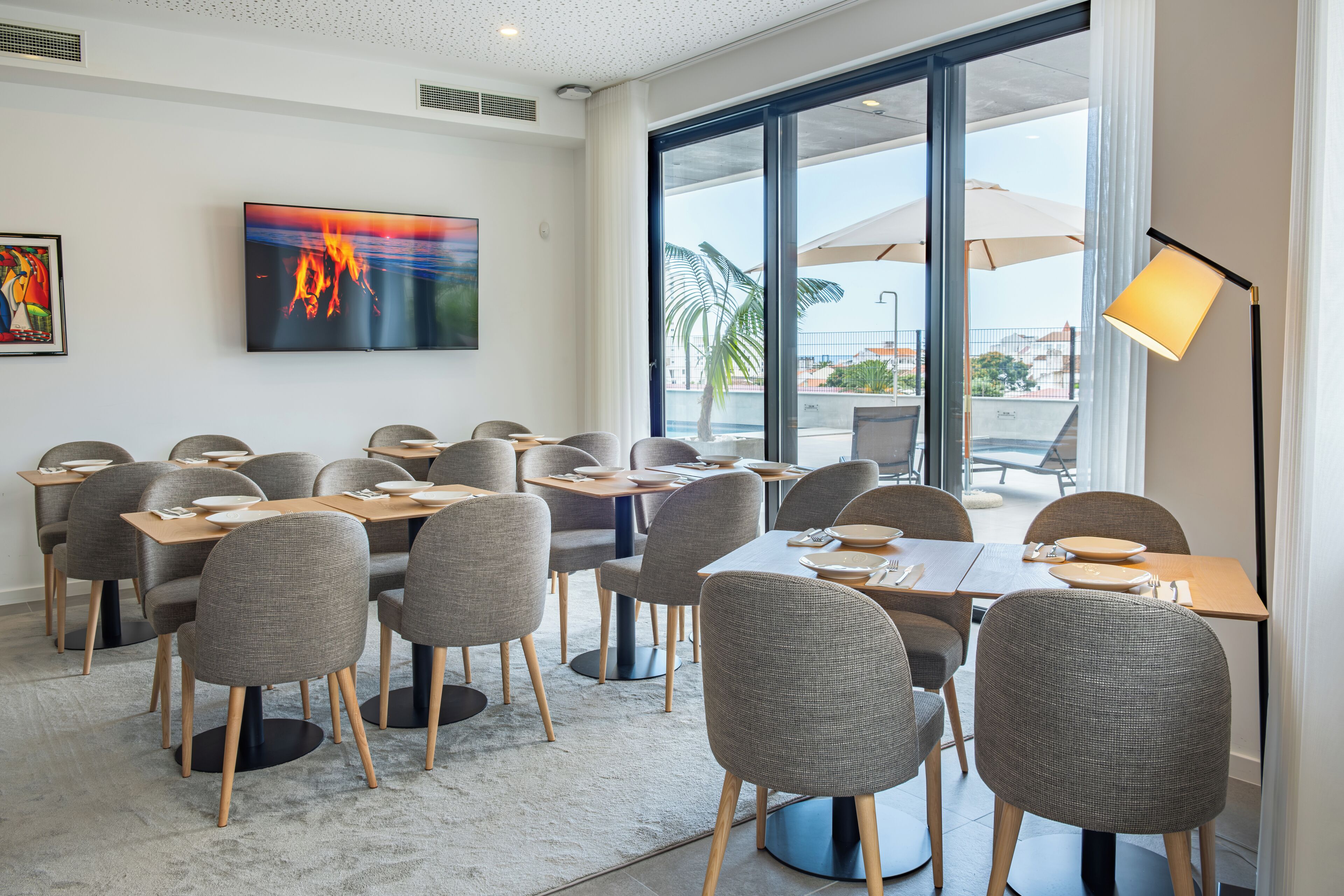 A modern dining area with grey upholstered chairs and tables, a large TV on the wall, and a view of the outdoors through large windows.
