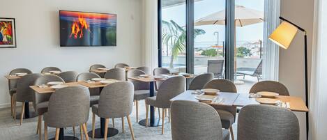 A modern dining area with grey upholstered chairs and tables, a large TV on the wall, and a view of the outdoors through large windows.