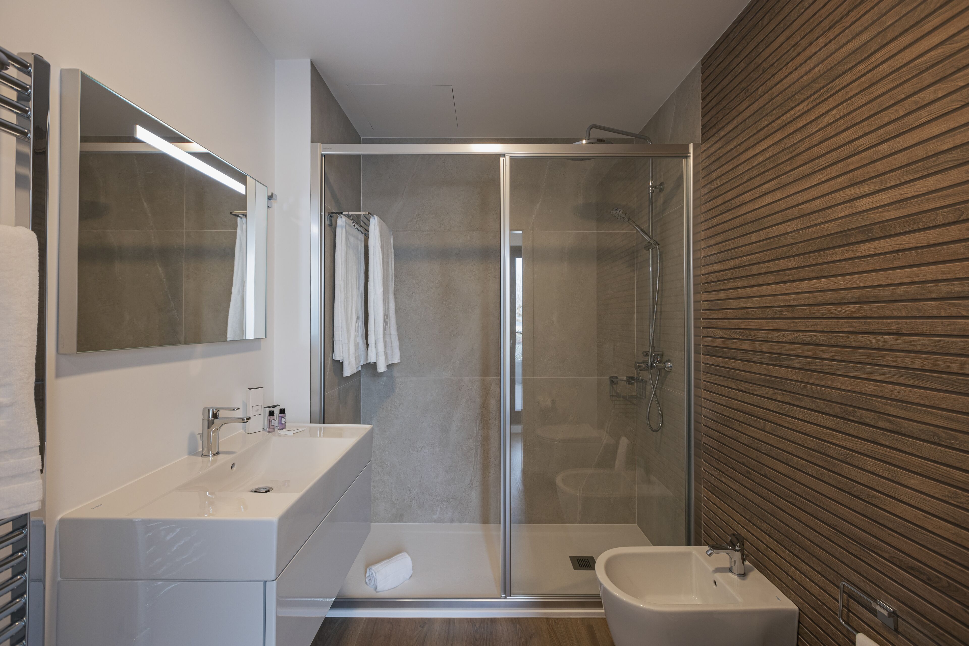 Une salle de bain moderne dotée d’une douche à paroi de verre, d’un meuble-lavabo à deux vasques et d’un mur revêtu de panneaux en bois.