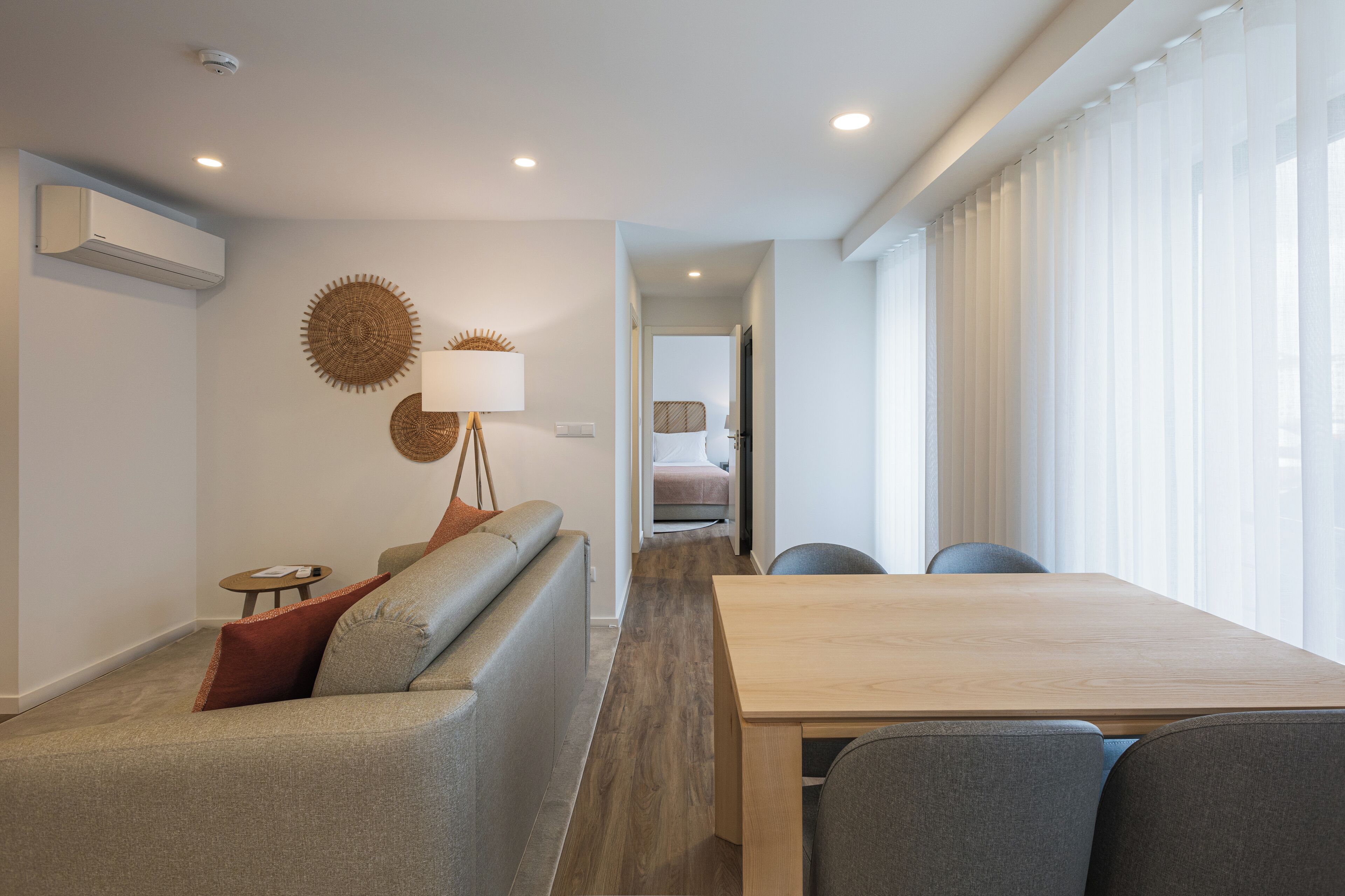 A modern hotel room with a sofa, a dining table, and a view of the city through large windows.