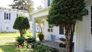 A white house with a covered porch, a small garden with various plants and flowers, and a tree in front of the house. - Spacious Private Guest Suite near Colgate (Earlville)