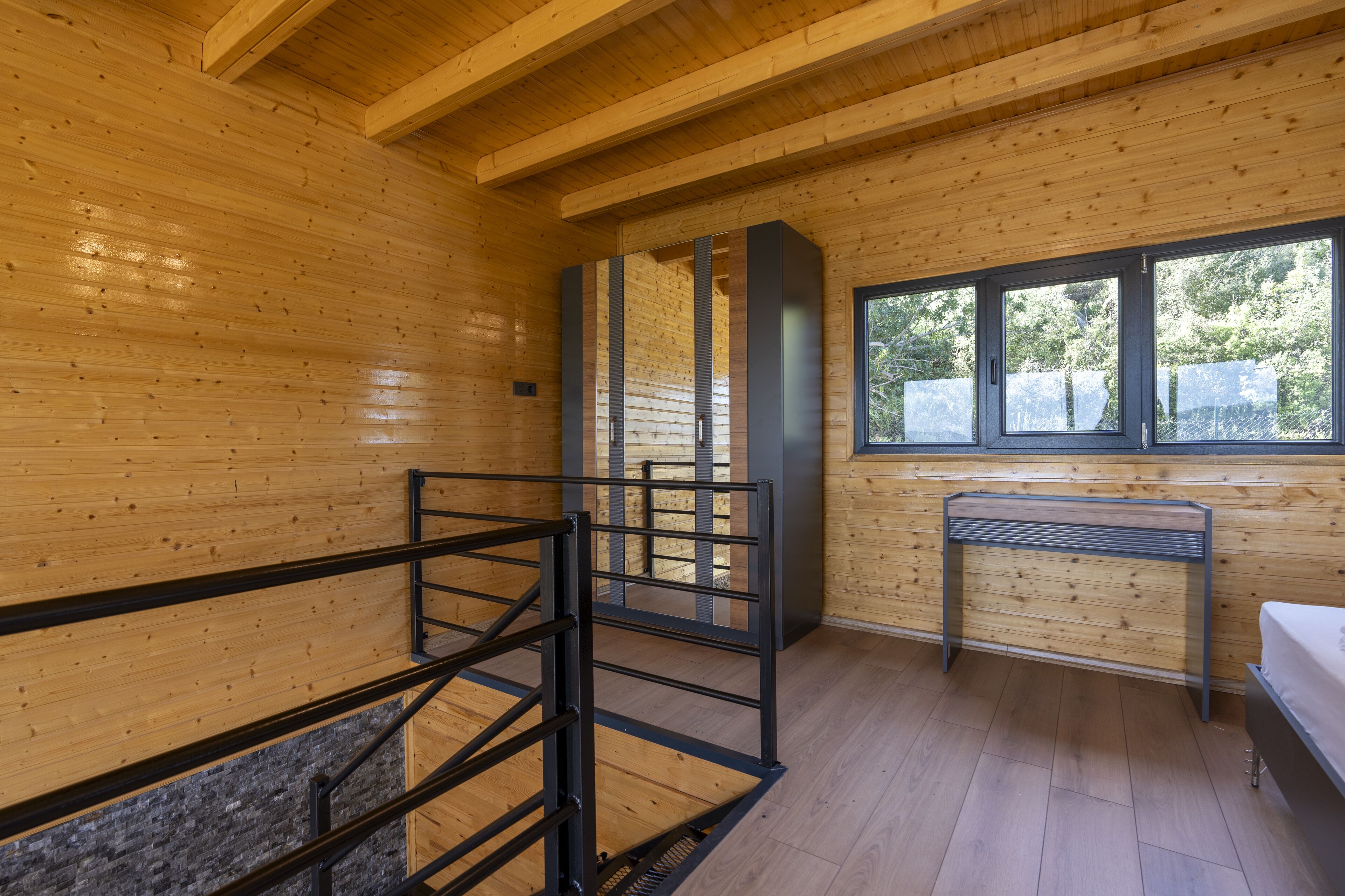 A wooden cabin interior with a metal staircase, a bench, and a window with a view of trees.