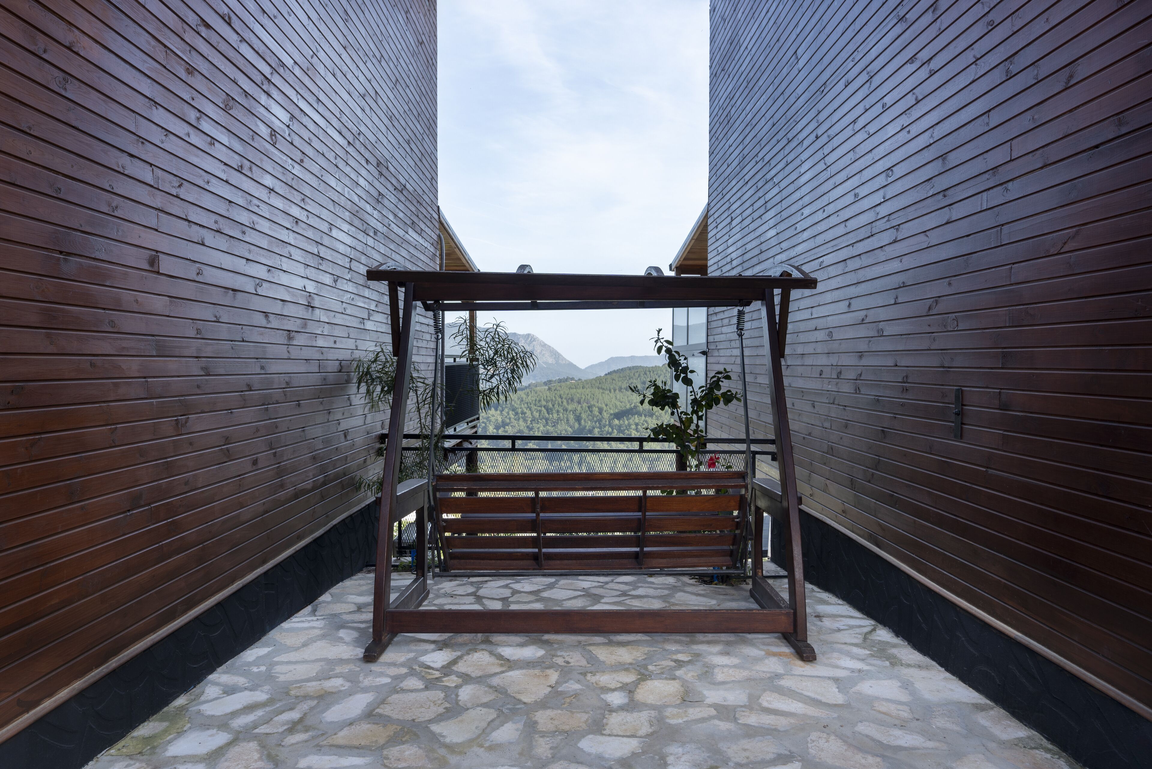 Un banco de madera con estructura metálica en un estrecho pasillo con suelo de piedra y vistas a montañas.