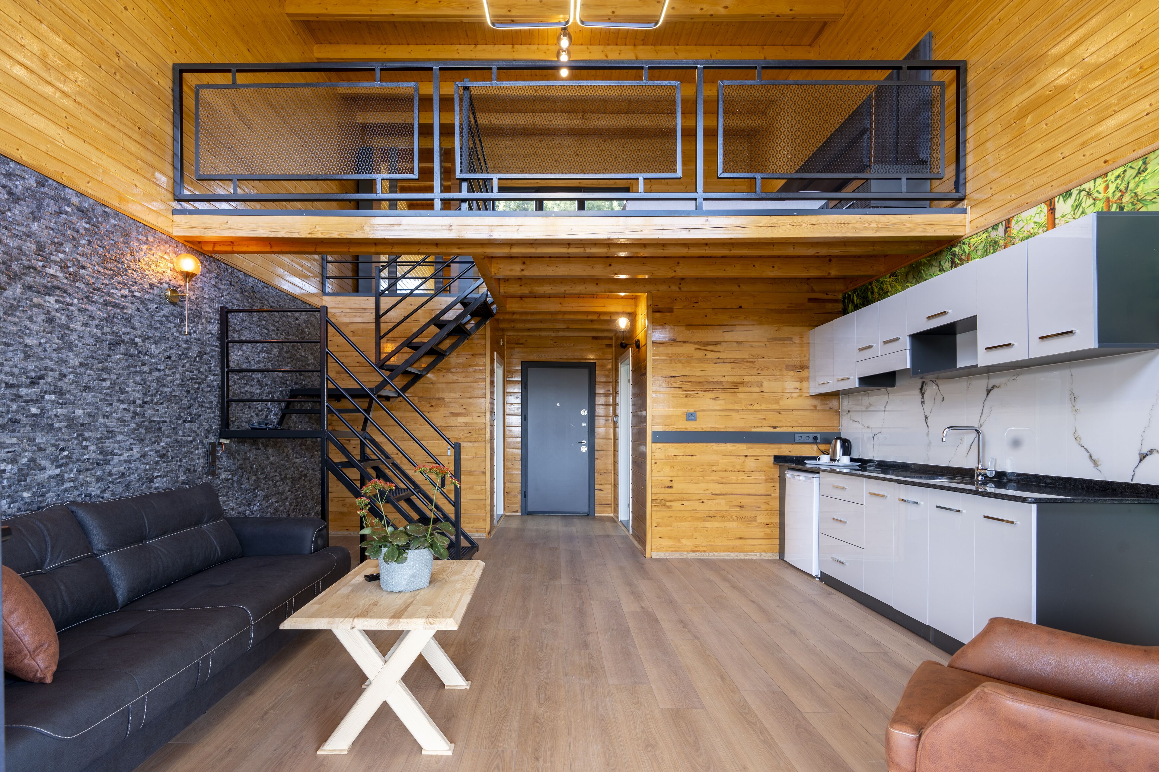 A modern interior with a wooden floor, black leather sofa, a wooden table, a staircase, and a kitchen area.