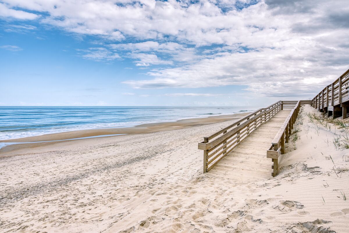 Ein Holzsteg führt zu einem Strand mit blauem Himmel und ruhiger Wellenbewegung.