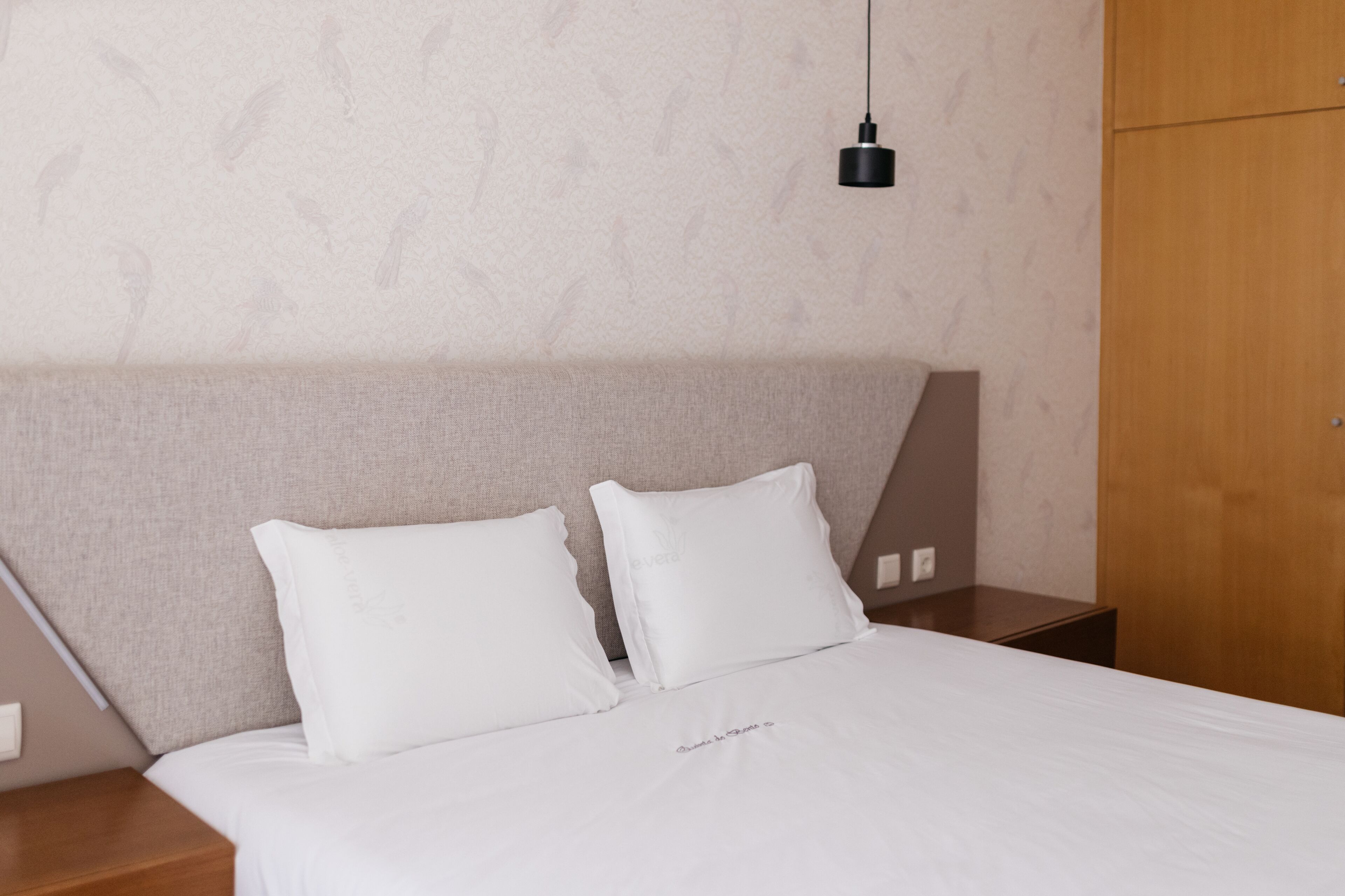 A neatly made bed with white bedding and two pillows, a bedside table, a hanging light fixture, and a wooden wardrobe.