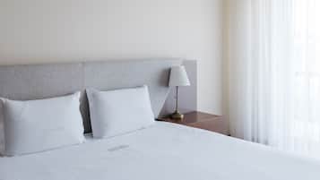 A neatly made bed with white bedding and pillows, a bedside lamp, and a window with sheer curtains.
