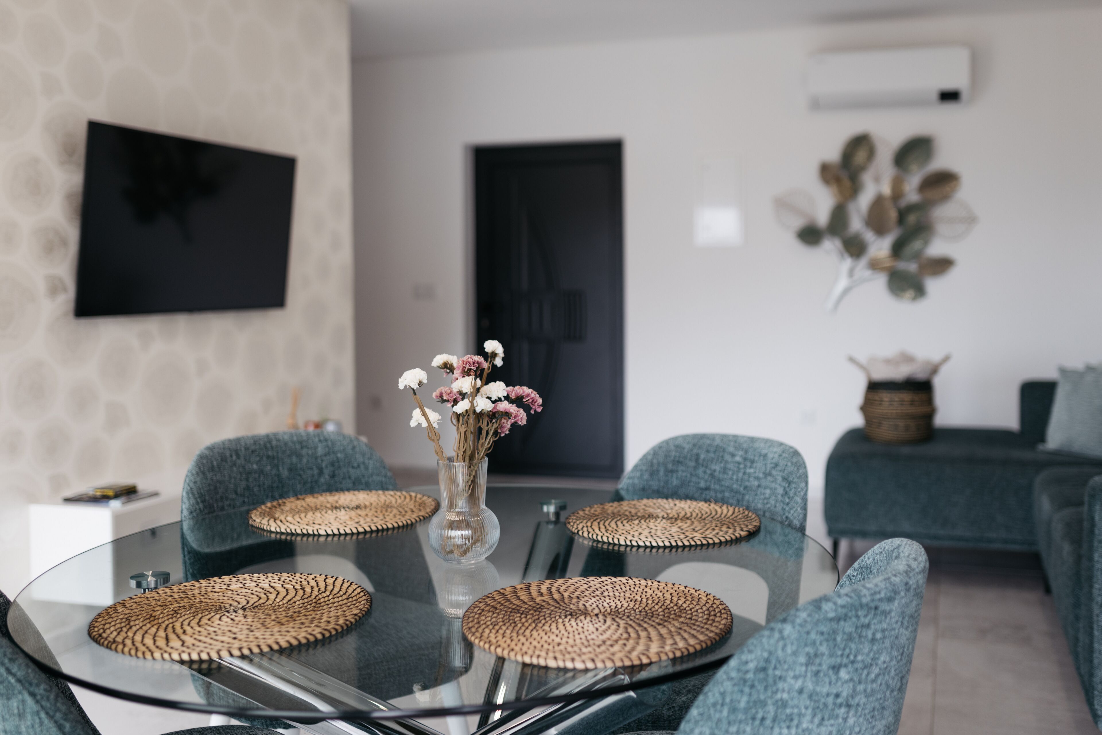 A modern dining area with a glass table surrounded by four chairs, a vase with flowers, and a decorative wall piece.