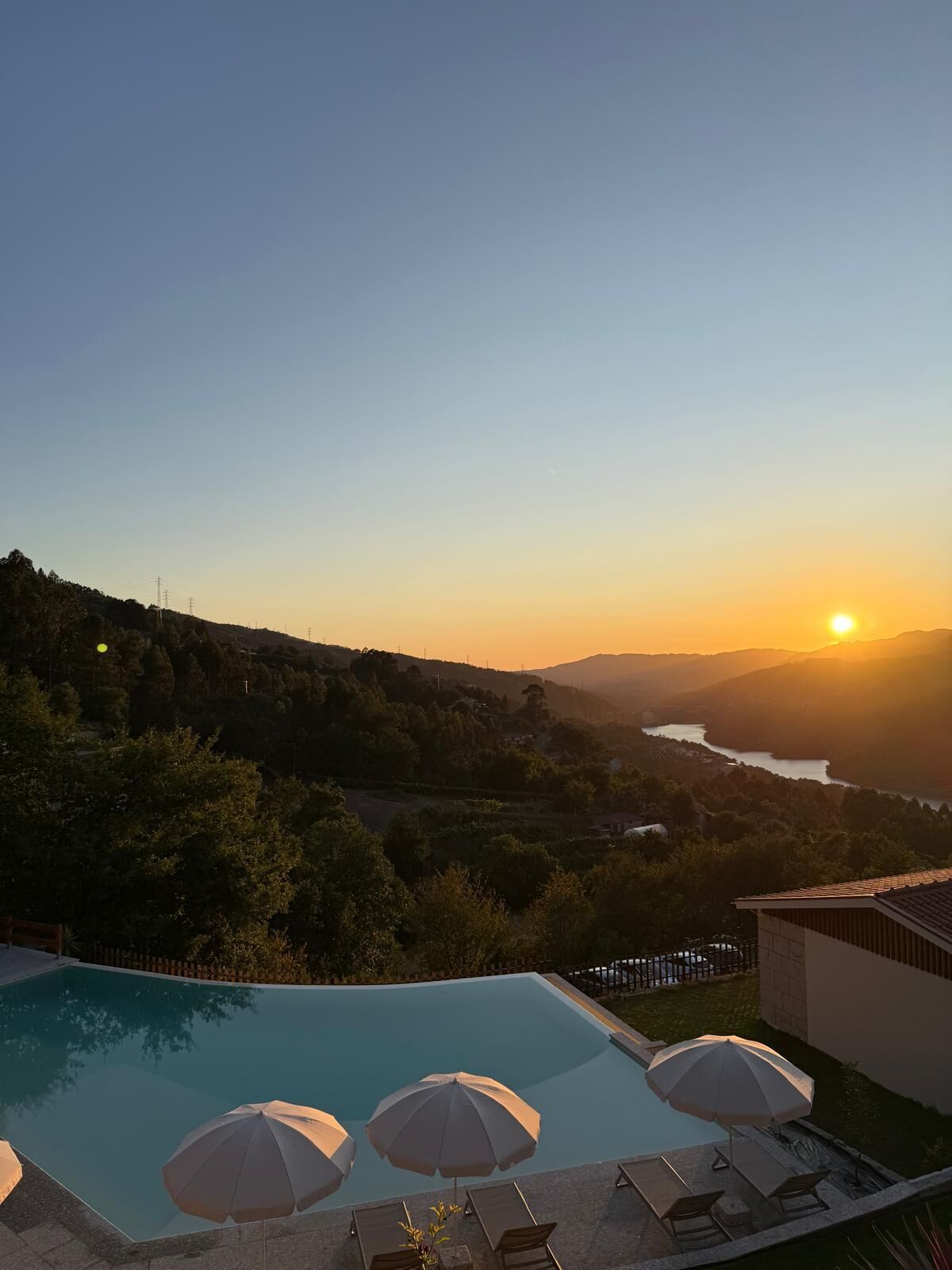 A swimming pool with lounge chairs and umbrellas, surrounded by a hilly landscape and a sunset view.