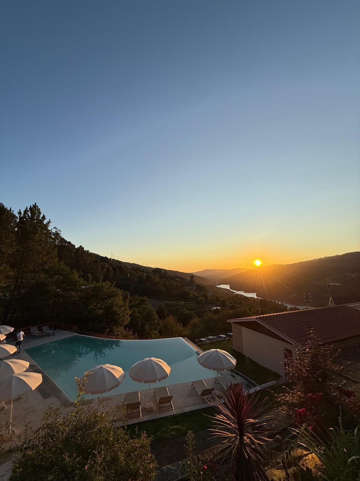 Une piscine entourée de verdure, dotée de chaises longues et de parasols, et offrant une vue sur une vallée où l’on peut admirer le coucher du soleil.