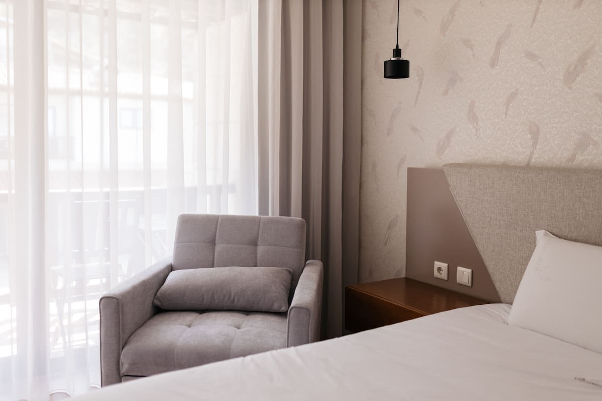 Una habitación de hotel moderna con una cama, un sillón gris, una mesita de noche y una lámpara colgante.