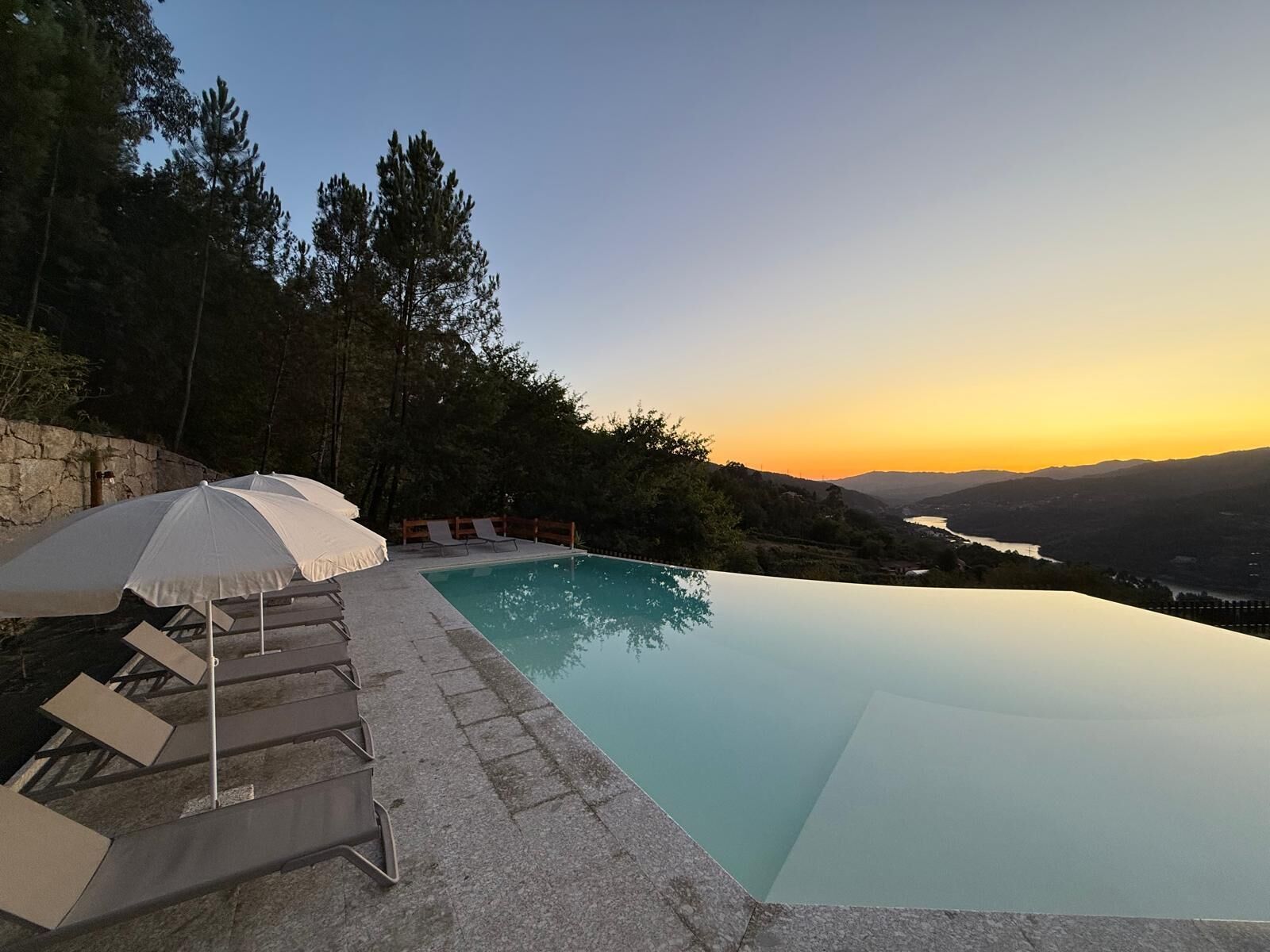 Una piscina infinita con vistas a la puesta de sol sobre un valle, rodeada de árboles y un muro de piedra.