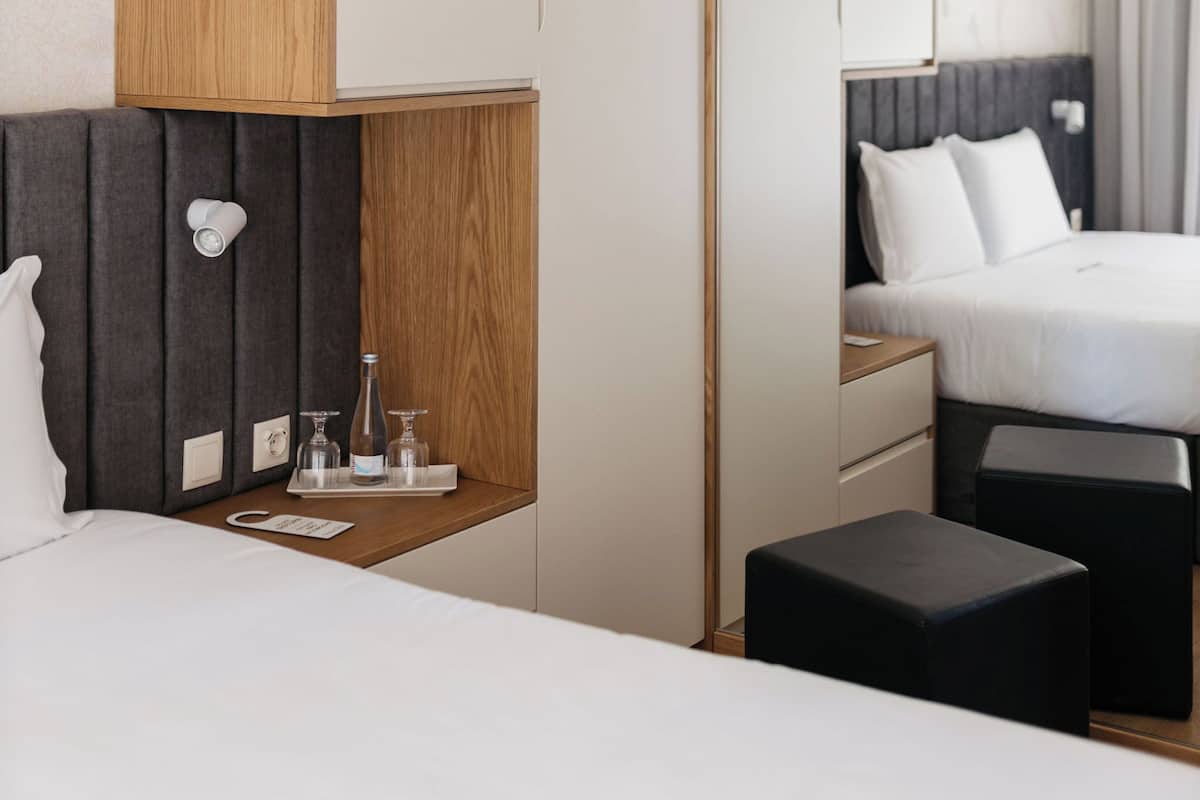 Una habitación de hotel moderna con un armario de madera, una mesita de noche con una bandeja, una cama y un pequeño otoman negro.