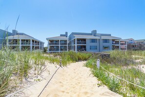 A beachfront property with a sandy pathway leading to a building with multiple balconies. - Oceanfront condo in Nags Head w/ community pool just steps to the sand! (South Nags Head)