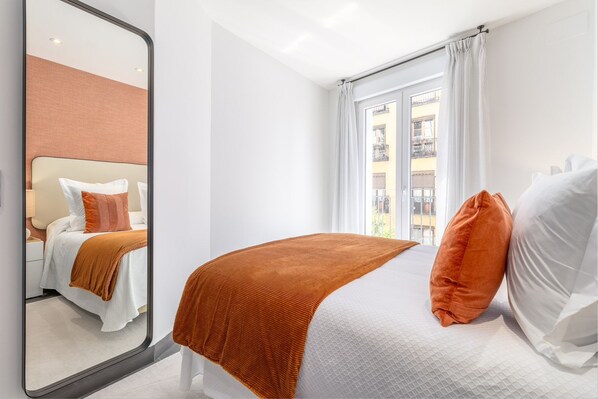 Ein Hotelzimmer mit einem großen Bett, einem Spiegel, einem Fenster mit Vorhängen und Blick auf Gebäude.