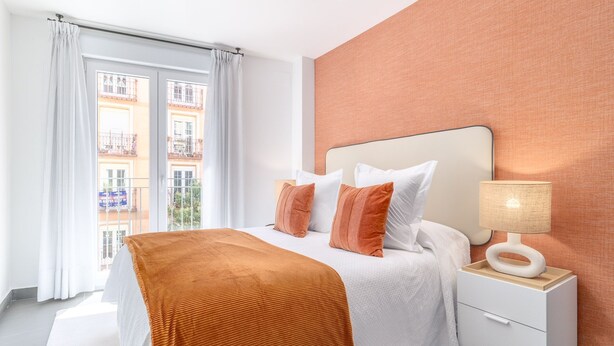 Ett modernt hotellrum med en stor säng, vita sängkläder, en träpanel som huvudgavel, ett nattduksbord med en lampa och utsikt över byggnader genom fönstret.