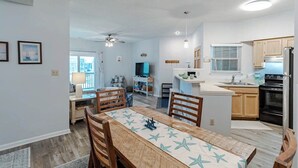 A modern kitchen with wooden cabinets, a black refrigerator, and a dining area with a table and chairs. - Great Coastal Condo in the heart of Kill Devil Hills NC! (Kill Devil Hills)