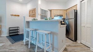 A modern kitchen with a central island, bar stools, wooden cabinets, a stainless steel refrigerator, and a blue accent wall. - Great Coastal Condo in the heart of Kill Devil Hills NC! (Kill Devil Hills)