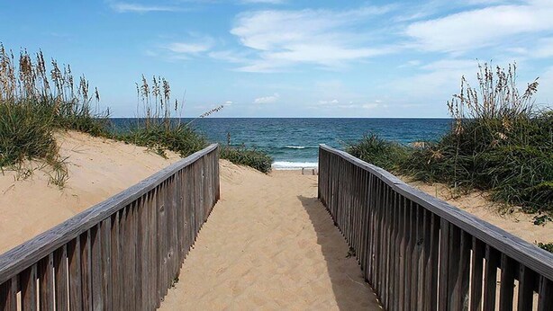 A wooden boardwalk leading to the beach with sand and vegetation on either side.
