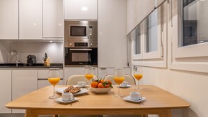 Une cuisine moderne équipée d’un four à micro-ondes, d’un four et d’une machine à café. On y trouve une table à manger avec des chaises, un saladier rempli de fruits et des verres de jus d’orange.