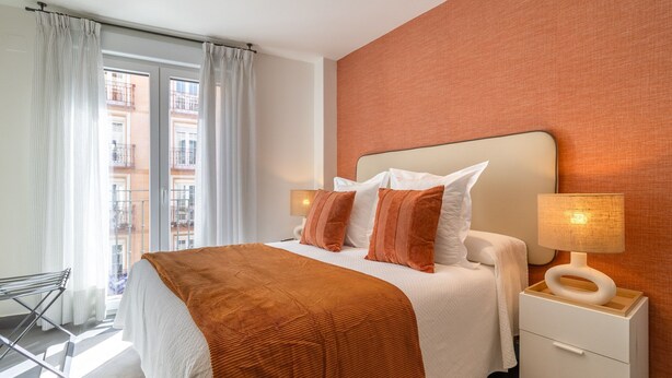 Une chambre avec un grand lit, une table de chevet avec une lampe et une vue sur les immeubles par la fenêtre.