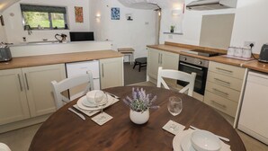 A cozy kitchen with a wooden dining table set for two, white cabinets, and a window overlooking greenery. - Le Durkestrete Cellar (Haverfordwest)