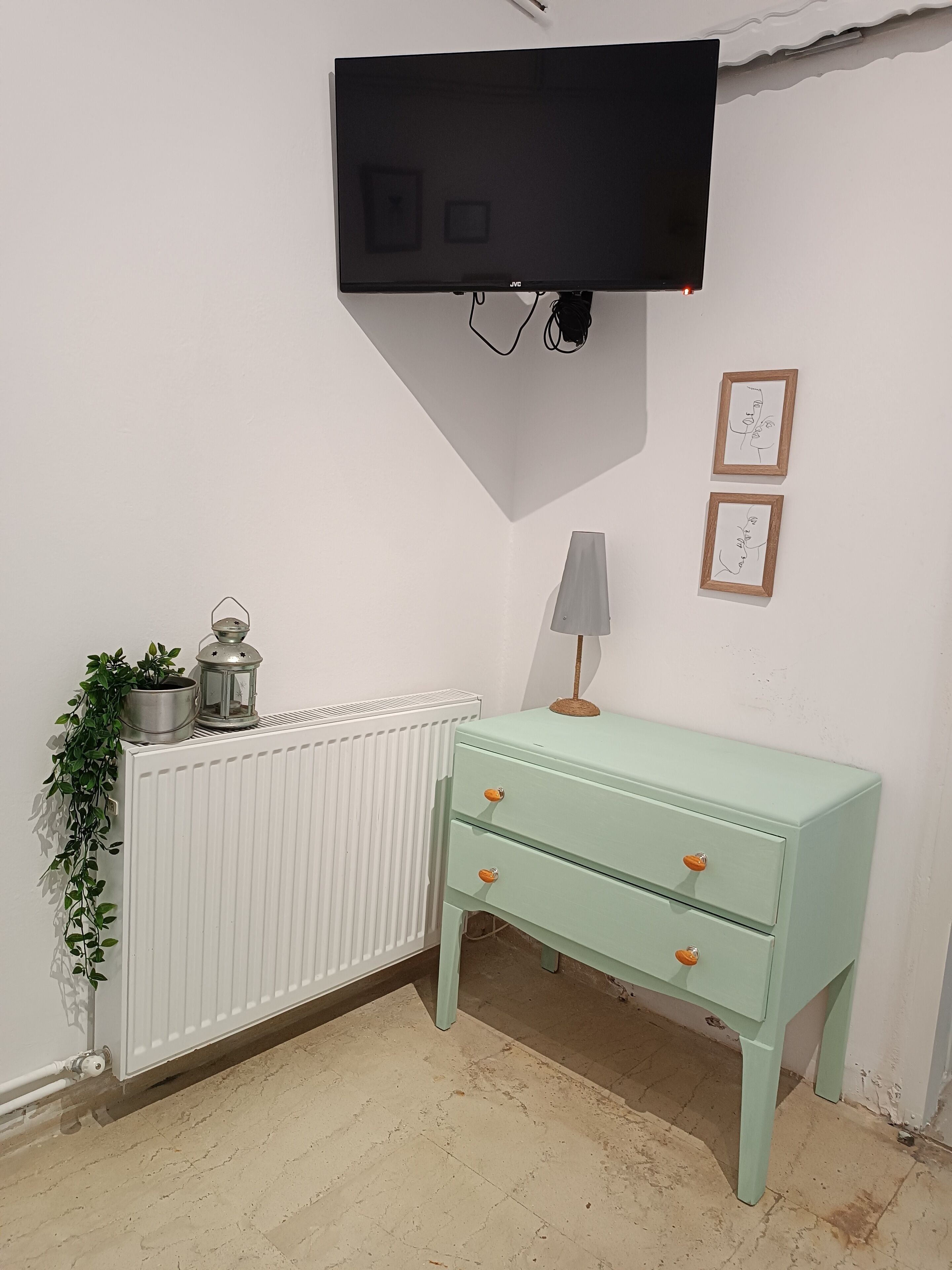 A room with a white radiator, a green dresser with drawers, a wall-mounted TV, and two framed pictures on the wall.