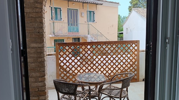 Un balcon ouvert, agrémenté de meubles en osier, d’une table en verre et offrant une vue sur un bâtiment doté d’un toit en tuiles et de volets verts.